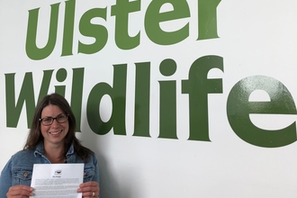 Annika Clements holding the Sustainable Fish Cities NI Pledge in front of a large Ulster Wildlife sign