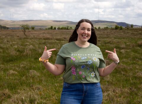 Model wearing Ulster Wildlife women's wildflowers t-shirt in green colour, pointing at t-shirt with each hand